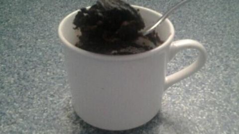 Ein weißer Becher mit einem schokoladigen, veganen Mikrowellen-Brownie steht auf einer blauen Arbeitsplatte.