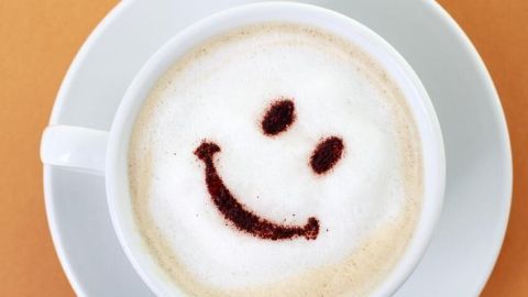 Eine Tasse Kaffee zeigt einen Smiley im Milchschaum, platziert auf einem weißen Unterteller, vor einem hellbraunen Hintergrund.