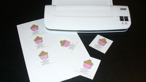 Laminiergerät verarbeitet Visitenkarten mit bunten Muffin-Designs auf Tisch; Textdetails: "Name", "Wohnort", "Telefonnummer".