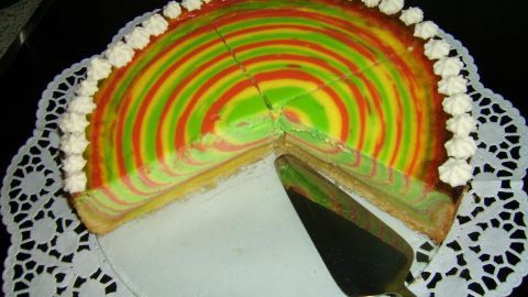Ein kunterbunter Quarkkuchen zeigt leuchtende Farbschichten, während ein Stück herausgeschnitten wird. Ein Serviermesser liegt bereit.