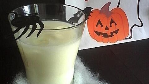 Ein Glas mit weißem Getränk, verziert mit einer Spinne, steht auf Watte. Im Hintergrund ein "BOO!"-Bild mit Geist und Kürbis.