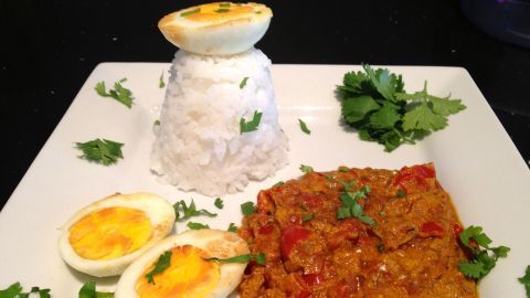 Auf dem Teller liegt ein vegetarisches Zwiebel-Curry mit gekochten Eiern und Reis, umgeben von frischen Kräutern.