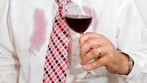 Ein Mann hält ein Glas Rotwein, während seine weiße Hemdbrust mit Flecken in einem hellen Raum markiert ist.