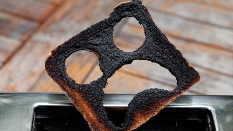 Ein verbranntes Toastbrot steckt in einem Toaster, umgeben von Holzboden; Acrylamid entsteht beim starken Rösten.
