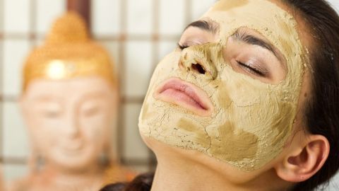Eine Frau mit einer Heilerde-Maske entspannt im Spa-Umfeld, während eine Buddha-Statue im Hintergrund sichtbar ist.