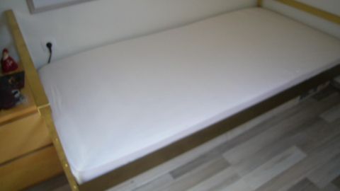 Ein Bett mit einer weißen Matratze steht in einem Raum. Der Raum hat einen Holzboden und eine Holzkommode.