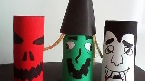 Drei gruselige Halloween Figuren aus Papierrollen stehen auf einem Tisch: ein rotes mit Schädelgesicht, ein grüner Monsterkopf mit Hut und ein schwarzes Vampirgesicht.