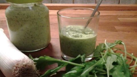 Ein Glas mit Pesto Verde steht auf einem Tisch, umgeben von frischen Gemüseresten wie Rucola und Lauch.