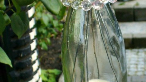 Ein hängendes Windlicht aus einer Flasche mit bunten Perlen und einer Kerze, hängt in einem Garten zwischen Pflanzen.
