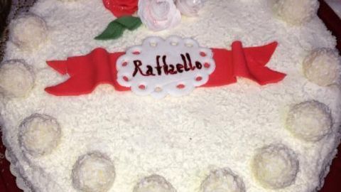 Eine Raffaello Torte mit weißen Kugeln und roten Zuckerdekoren thront auf einem Tisch, verziert mit "Raffaello" Schriftzug.