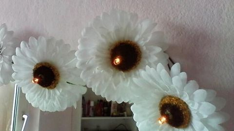 Eine Lichterkette mit großen, weißen Blüten leuchtet sanft und schmückt eine Wand in einem gemütlichen Raum.