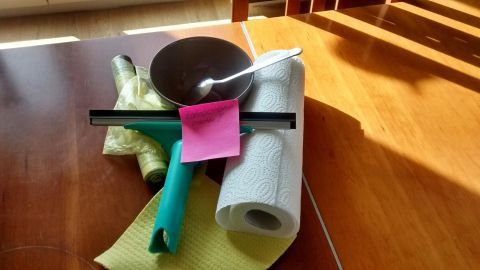 Ein Teller und Besteck liegen auf einem Holztisch, umgeben von Putzmaterialien, ideal für den ersten Haushalt.