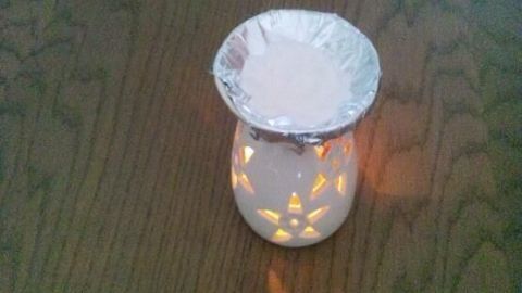 Ein weißer Keramikduftlampe leuchtet warm auf einem Holztisch; Vanillezucker liegt in Alufolie auf der Lampe.