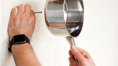 Eine Hand hält einen Kochtopf, der als Hammerersatz verwendet wird, um einen Nagel in die Wand zu treiben.