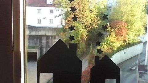 Schwarze Fensterdekoration in Form von Häusern und Sternen ziert eine Fensterscheibe, vor der bunte Herbstbäume stehen.