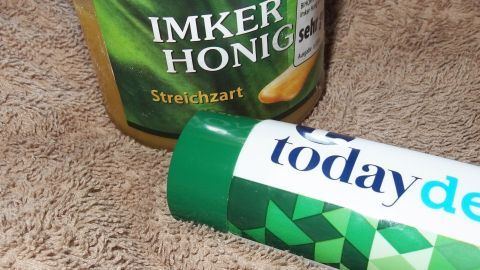 Ein Glas „Imker Honig“ und eine Tube Zahnpasta mit der Aufschrift „today“ liegen auf einem beigen Handtuch.
