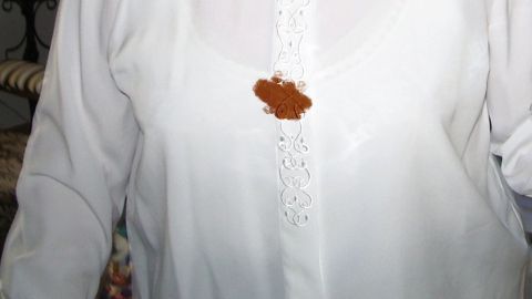Eine weiße Bluse mit einem braunen Schokoladeneis-Fleck in der Mitte, auf einem neutralen Hintergrund.