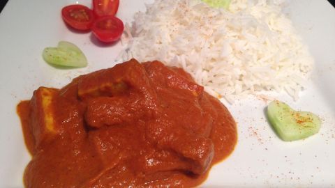 Ein Teller zeigt "Butter Chicken" mit gebratenem Tofu und Beilage aus Reis, garniert mit Kirschtomaten und Gurken.