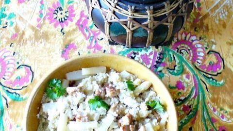 Ein bunter Teller mit Couscous-Salat, gemischt mit Huhn und Feta, steht auf einem dekorativen Tuch. Daneben ein stilvoller Behälter.
