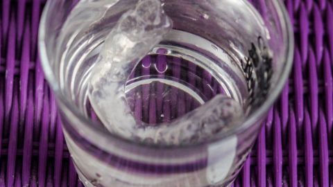 Ein Glas Wasser mit einer sprudelnden Reinigungstablette für Aufbissschiene/Knirscherschiene auf einer lila Mattenoberfläche.