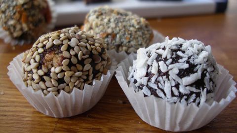 Leckere Schoko-Keks-Pralinen liegen in Papierförmchen auf einem Holztisch und sind mit Sesam und Kokos bestreut.