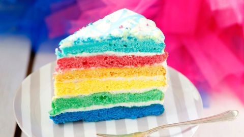 Ein Stück Regenbogenkuchen mit leuchtend bunten Schichten liegt auf einem Teller, die Schichten von Rot bis Blau.