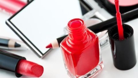Roter Nagellack steht geöffnet auf einem Tisch umgeben von weiteren Kosmetika wie Lippenstift und Kajalstiften.