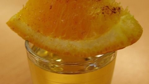 Ein Glas mit gelbem Getränk steht auf einem Tisch, gekrönt von einer orangefarbenen Scheibe, die gewürzt ist.
