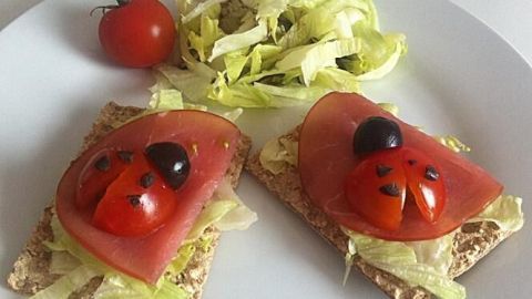 Zwei Brotscheiben sind mit Salat und Tomaten belegt, die wie Marienkäfer aussehen, umgeben von einem kleinen Salat und einer Tomate.