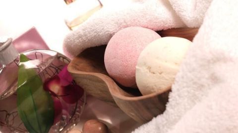 Badekugeln liegen in einer Holzschale, umgeben von weichen Handtüchern mit Parfümflaschen und Dekosteinen im Hintergrund.