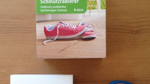 Schmutzradierer liegt neben einer Schere auf einem Holztisch, die Verpackung zeigt ein Paar rote Schuhe mit Schnürsenkeln.