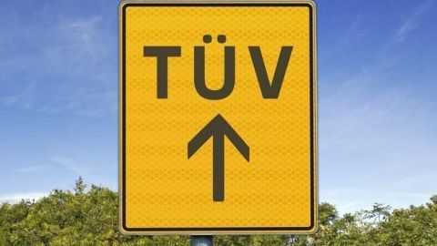 Ein gelbes Schild zeigt "TÜV" mit einem Pfeil nach oben, im Hintergrund blauer Himmel und Büsche.