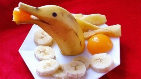 Ein Delfin aus einer Banane hält ein Stück Obst und steht auf einem Teller, umgeben von Bananenscheiben und einer Gelbe.