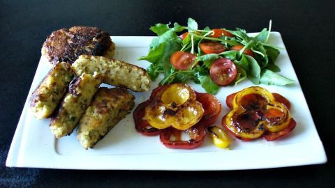 Bulgur-Bratlinge liegen auf einem weißen Teller neben frischem Salat und Tomatenscheiben in einer dunkel gefärbten Umgebung.