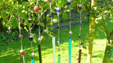 Ein farbenfrohes Windspiel hängt dekorativ an einem Baum im Garten und reflektiert Sonnenlicht, umgeben von grünen Blättern.