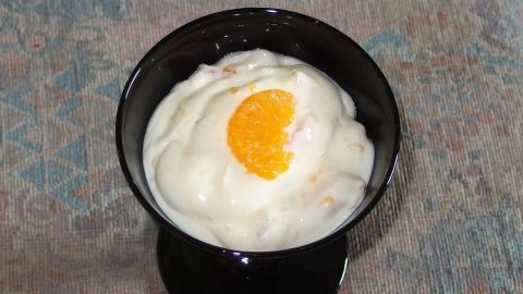 Ein schwarzes Dessertglas enthält cremigen, käsekuchenartigen Nachtisch, garniert mit einer halben Orange. Der Hintergrund zeigt eine strukturierte Oberfläche.