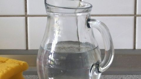 Eine Glas Umschüttkanne füllt sich mit Wasser auf einer Küchentheke, neben einem gelben Schwamm und Fliesen.