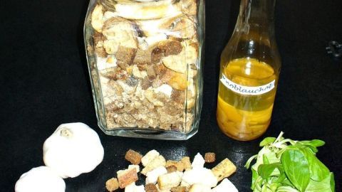 Ein Glas mit Croutons steht neben Knoblauch, Öl und frischem Gemüse auf einem dunklen Tisch, bereit zur Zubereitung.