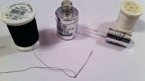 Ein schwarzer und ein heller Nähfaden liegen neben einer Nadel, während der Nähfaden in die Nadel eingefädelt wird.