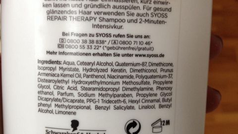 Ein Shampoo-Flasche steht im Fokus, umgeben von einem Holzhintergrund. Auf dem Etikett steht, dass der Syoss-Stylist empfiehlt, das Produkt zu verwenden.