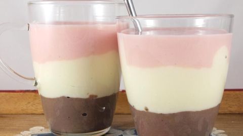 Zwei Glasbecher mit einem geschichteten Eiweißshake in verschiedenen Farben, stehen auf einem blumigen Tischdeckenmuster.