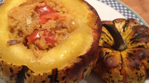 Ein halbierter Gorgonzolakürbis mit goldbrauner Couscous-Füllung und Gemüse steht auf einem Teller neben einem ganzen Kürbis.