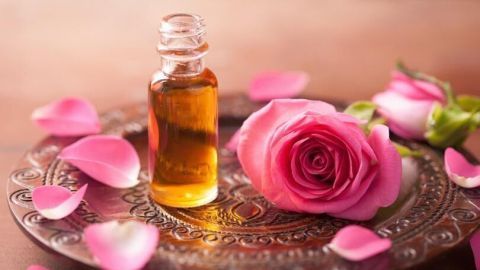 Ein Glasfläschchen mit goldenem Parfüm steht auf einem dekorativen Tablett neben einer rosa Blüte und Rosenblättern.