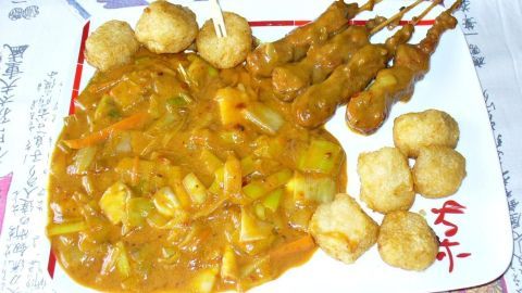 Eine bunte Lauch-Gemüsepfanne mit Erdnusssoße liegt auf einem Teller, daneben leckere Erdnussspieße und frittierte Bällchen.