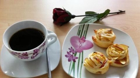 Eine Tasse Kaffee steht neben einem Teller mit Apfel-Blätterteig-Rosen, dekoriert mit einer roten Rose auf dem Tisch.