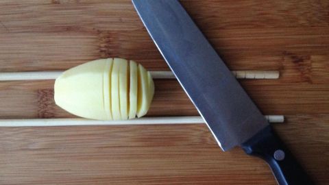 Eine Kartoffel mit gleichmäßigen Schnitten liegt zwischen zwei Essstäbchen auf einem Holzbrett, während ein scharfer Messer daneben liegt.
