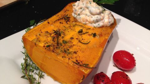 Ein gebackener Butternut-Kürbis liegt auf einem Teller, garniert mit Kräutercreme und roten Kirschtomaten.