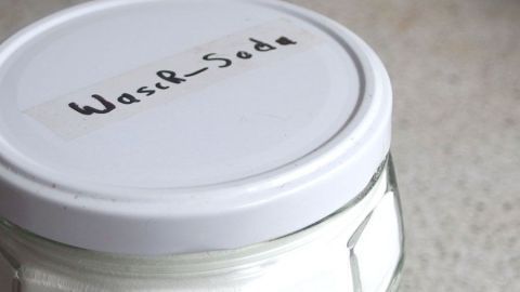 Ein Glasbehälter mit einem weißen Deckel steht auf einer stark verschmutzten Küchenarbeitsplatte aus Kunststoff. Auf dem Deckel steht "Waschsoda" geschrieben.