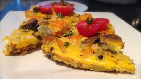 Ein goldgelbes Omelett mit Butternut-Kürbis und Champignons liegt auf einem weißen Teller, dekoriert mit Kirschtomaten.