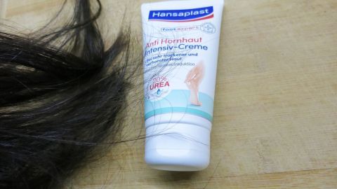 Fußcreme für gesunde Haare? Entdecke, wie die intensive Pflege mit Urea auch deinem Haar Glanz und Geschmeidigkeit verleiht. Ein überraschender Tipp für geschmeidige Mähnen! Eine Tube Hansaplast Anti Hornhaut Intensiv-Creme liegt neben dunklem Haar auf einer Holzoberfläche. Text: "20% UREA".